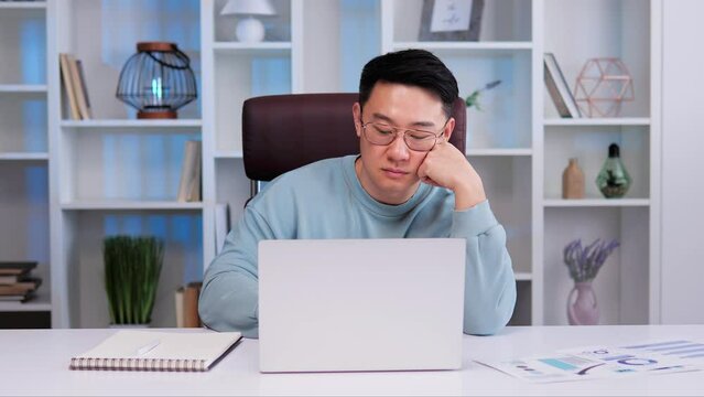 Asian male with glasses is sitting at desk in the office. Male appears bored, sad, exhausted tries to work on his laptop. He seems to have no motivation or interest in his work, experiencing burnout.