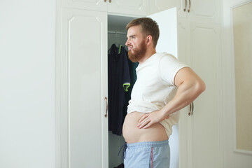 Fototapeta na wymiar Unhappy sad upset young man with fat on her stomach, guy worried about body fat touching belly standing at home near closet
