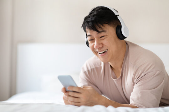 Positive asian man using smartphone and headphones in bed