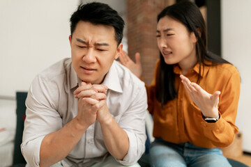 Fototapeta na wymiar Angry asian lady shouting at her middle aged husband, having conflict while sitting on sofa at home, focus on upset man