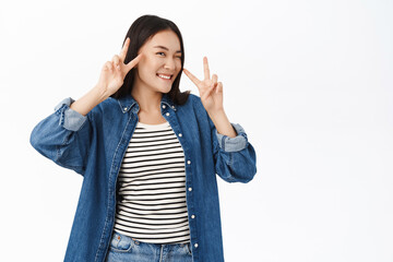 Happy asian girl shows peace, v-signs near face and smiles at camera, positive face expression, stands over white background