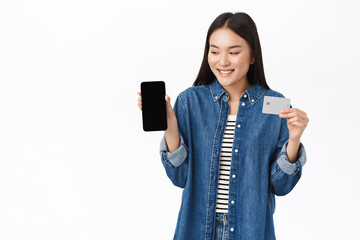 Smiling asian girl shows smartphone screen and credit card, mobile banking, standing over white studio background