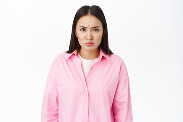 Portrait of angry asian girl frowns, looks offended at you, negative emotions, stands over white...