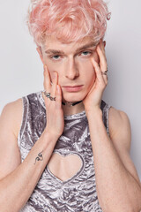 Serious pink haired man with non traditional orientation touches face looks directly at camera wears stylish t shirt and rings on fingers looks attentively at camera poses against white background.