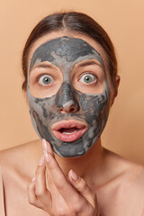 Headshot of shocked stunned woman with dark combed hair gets beauty treatments applies facial clay mask holds breath from amazement poses indoor takes care of skin isolated over brown background