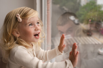 Blonde two year old girl at home looking through window at street.Copy space. 