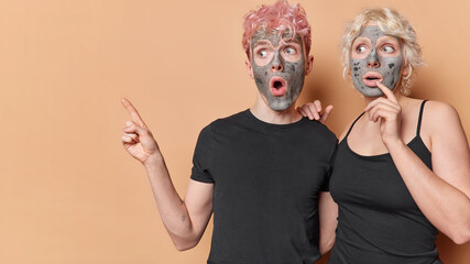 Studio shot of surprised shocked boyfriend and girlfriend wear casual black t shirts apply facial mud mask for reducing pores and blackhead advertise beauty product point away on blank space
