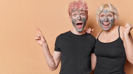 Crazy emotional woman and man advertise something awesome point aside on left side apply facial clay mask pose against brown background undergo beauty procedures. Wellness and skin care concept