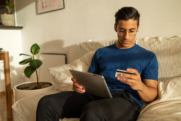 Attractive Hispanic man with glasses wearing blue shirt in the apartment using tablet and looking at the credit card