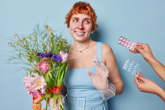 Happy redhead woman refuses to take medicine holds bouquet of flowers suffers from pollen allegry smiles toothily wears dress and jewellery isolated over blue background suffers from runny nose