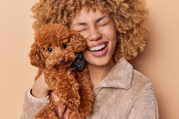 Photo of beautiful cheerful curly haired female pet owner smiles happily holds small adorable poodle puppy near face likes animals smiles broadly wears fur coat isolated over brown background.