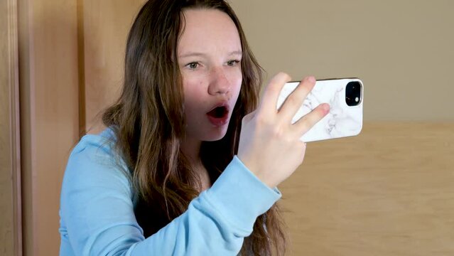 girl holds phone horizontally she is very surprised, she opens mouth strongly looking at herself in reflection in mirror surprise to be interested in changes in appearance pimple on face
