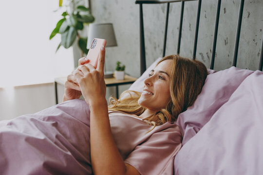 Young Woman Wear Purple T-shirt Pajama Lying In Bed Hold In Hand Use Mobile Cell Phone Resting Relax Spend Time In Bedroom Lounge Home In Own Room Hotel Wake Up Dream Be Lost In Reverie Good Mood Day