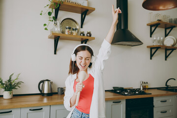 Young cheerful singer fun positive caucasian housewife woman wearing casual clothes headphones listen music use spoon as microphone cooking food in light kitchen at home alone. Healthy diet concept.