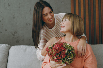 Two fun adult women mature mom young kid wear casual clothes give bouquet of hydrangea flowers sit on gray sofa couch stay at home flat rest relax spend free spare time in living room. Family concept.