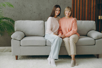 Full body two adult women mature mom young kid wear casual clothes look camera hold hands hug sit on gray sofa couch stay at home flat rest relax spend free spare time in living room. Family concept.
