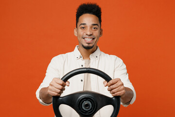Young smiling happy fun cheerful man of African American ethnicity wears light shirt casual clothes hold in hands steering wheel isolated on orange red background studio portrait. Lifestyle concept.