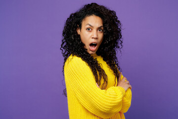 Side view young dissatisfied sad woman of African American ethnicity wearing casual yellow sweater hold hands crossed folded look camera isolated on plain purple background studio. Lifestyle concept.