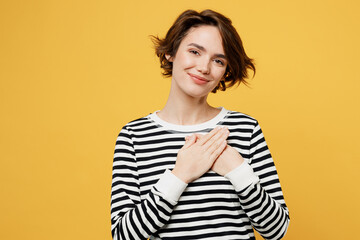 Young smiling grateful happy cheerful woman wear casual striped black and white shirt put folded hands on heart look camera isolated on plain yellow color background studio portrait Lifestyle concept
