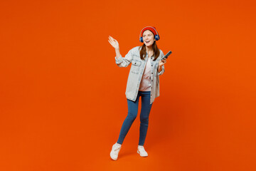 Full body young woman wear denim shirt white t-shirt red hat headphones listen to music use mobile cell phone look aside on area isolated on plain orange background studio portrait. Lifestyle concept.