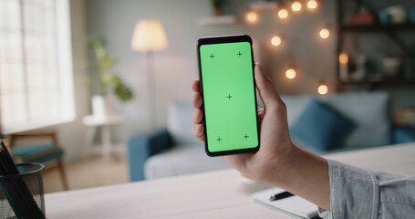 Close up shot of hand of guy holding a smartphone in cozy living room. A green screen mock up...
