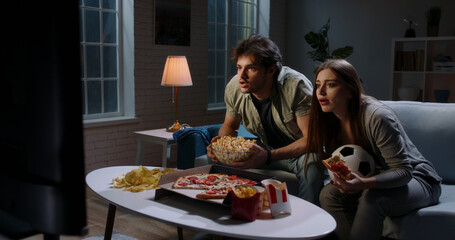 Funny young emotional caucasian sport fan couple watching a game together, eating popcorn and pizza and reacting to their team playing 