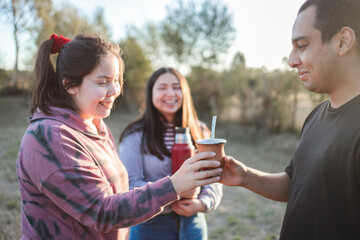  Group of smiling friends drinking yerba mate using a thermos with hot water in the countryside at sunset. - obrazy, fototapety, plakaty