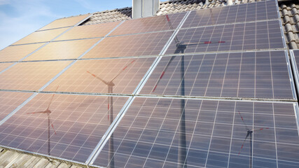 Solar power, photovoltaic panels of older construction on a house roof. Wind turbines and the sun are reflected in the panels.