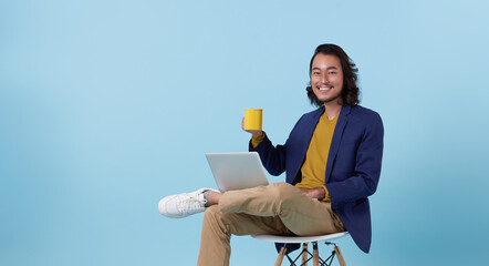 Young asian business man happy smiling using computer laptop sitting on white chair and drinking coffee isolated on blue studio background.
