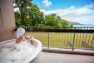 Young female in bath towel on head enjoys outdoor luxury spa, immersed in beautiful bath tub with sea view.