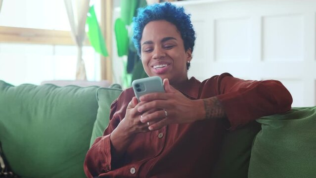 Young overjoyed African American woman with phone making victory gesture and shouting with happiness after receiving email with job offers with good salary and insurance sits on sofa in home interior