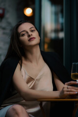 A stylish woman in a dress poses with a glass in her hand on the street of a restaurant. Vertical view