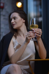 A stylish woman in a dress poses with a glass in her hand on the street of a restaurant. Vertical view