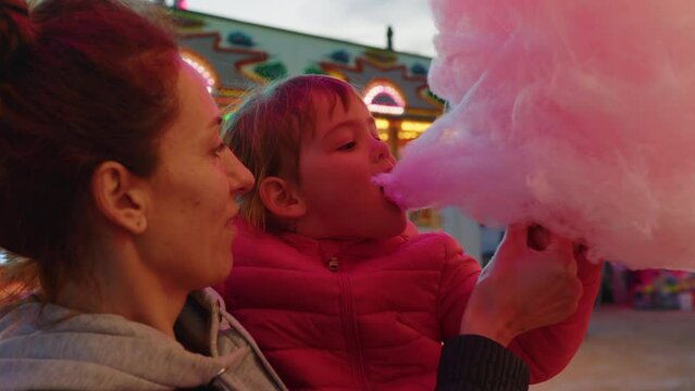 Close up shot of a happy smiling mother and his little daughter are having fun to eat together a cotton candy in amusement park with luna park lights at night