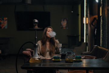 Beautiful young woman inhaling hookah girl smoking hookah sitting in lounge bar concept of...