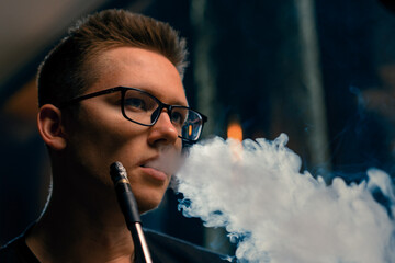 A man in glasses smokes a traditional hookah pipe A man exhales thick smoke in a hookah cafe or lounge bar - obrazy, fototapety, plakaty