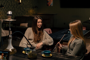 two young female friends are sitting relaxed in a hookah bar smoking hookah and having fun chatting...