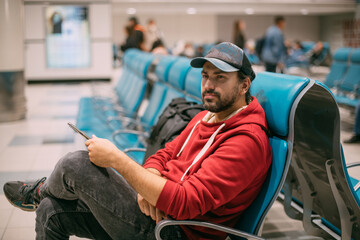 Fototapeta na wymiar A young, handsome man is sitting with a phone in his hands at the airport in the evening. A guy, a tourist with a smartphone and a backpack is waiting for departure at the gate
