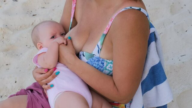 Cute baby girl sucking milk from breast of her mother on the beach. Family vacation and travel concept