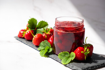 Strawberry confiture in glass jar on slate dish with fresh strawberry fruit and green leaves on marble. Recipe of delicious homemade berry jam of strawberry full of vitamins and antioxidants.