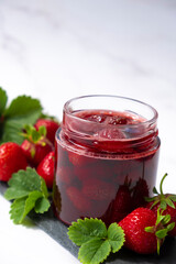 Homemade strawberry preserves or jam in glass jar on slate dish with fresh strawberry fruit and green leaves on marble. Recipe of delicious homemade berry jam of strawberry full of vitamins.