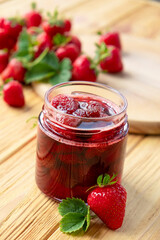 Strawberry jam in glass jar on wooden board with fresh strawberry fruit and green leaves on wooden background. Recipe of delicious homemade berry jam of strawberry full of vitamins and antioxidants.