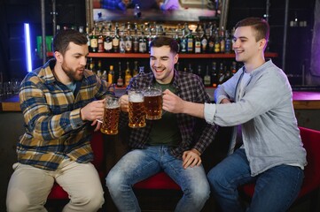 Obraz premium Three young men in casual clothes are smiling and clanging glasses of beer together while sitting at bar counter in pub