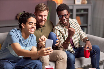 Group of three male friends watching sports match online via smartphone and cheering emotionally