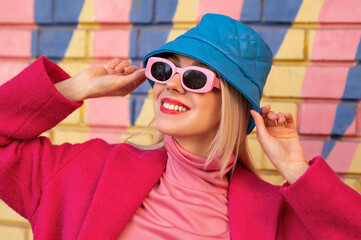 Fashionable happy smiling blonde woman wearing trendy pink sunglasses, turtleneck, blue leather...