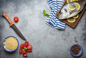 Raw fish dorado with ingredients lemon, fresh basil, cut cherry tomatoes, uncooked rice on wooden cutting board with knife on rustic stone background top view, cooking healthy fish dorado. Copy space