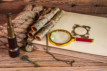 Old items, magnifying glass, map scrolls, pocket watch lying on an old book next to a rope, a telescope and an old key