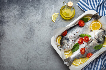 Raw fish dorado in casserole dish with lemon, fresh basil, cherry tomatoes, bottle of olive oil on wooden cutting board on rustic stone table top view, cooking healthy fish dorado. Copy space