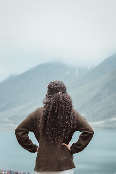 young curly hair woman in brown sweater hands on waist enjoying scenery fresh air clouds over the mountains in lake saif ul malook naran kaghan valley