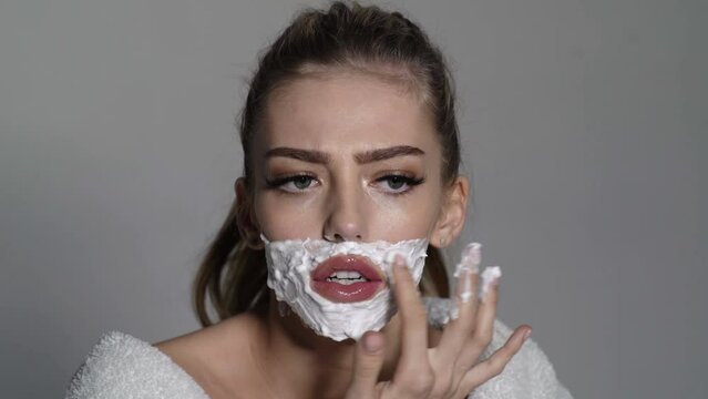 Woman with foam on face holds straight razor in hand. Girl on busy face wears bathrobe, grey background. Barber and shaving concept. Lady trans gender with sharp blade of straight razor.